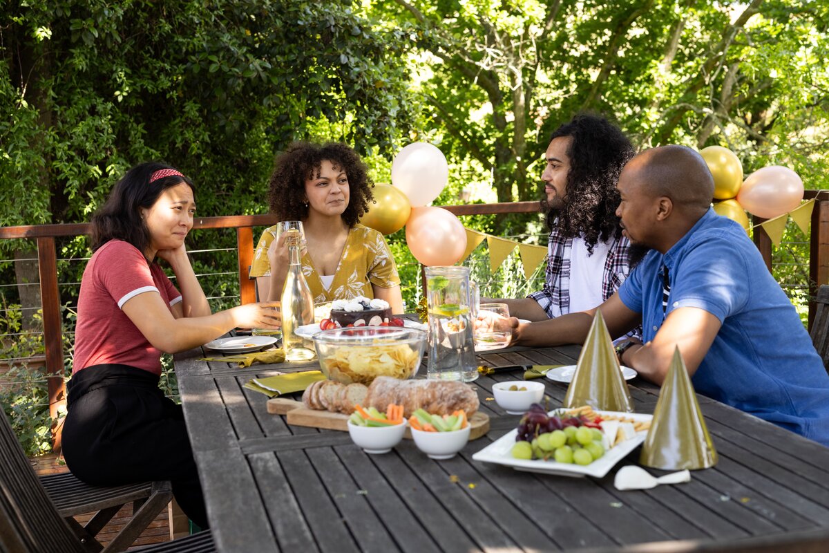 Guests seated at a wooden dinner table with balloons, cake and grazing platters at a backyard engagement party
